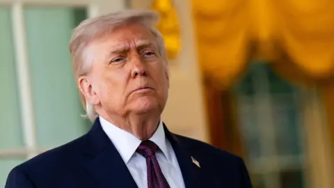 Donald Trump has a stern expression and wears a suit with gold behind