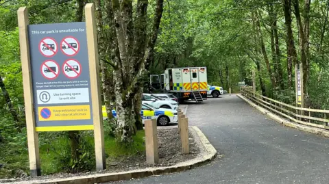 Coches de policía y una ambulancia aparcados en un aparcamiento rodeado de árboles al fondo. Un cartel en primer plano indica que el aparcamiento no es apto para ciertos vehículos grandes como remolques, camiones y autobuses.