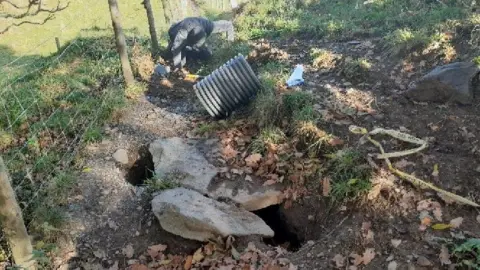 LDNPA Path which has several large holes after badgers dug tunnels underneath. There is a ranger working to repair the path and a large black pipe sits to the side. 