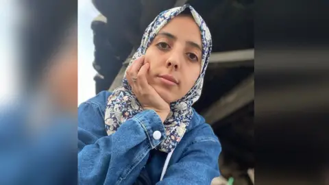 Asmaa Tayeh Selfie of Asmaa, whose head rests in her other hand. She has a serious expression on her face. She is wearing a blue denim jacket and a headscarf patterned with blue and white flowers. There appears to be a damaged building behind her