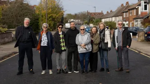 Eight residents stand in a row in the road with houses lining the side of the street. There are a mixture of men and women. The road features single yellow lines. 