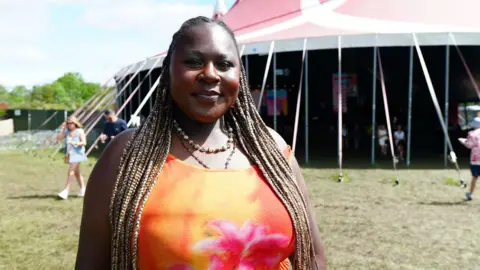 A woman with long hair styled in locs, wearing a blood-orange vest top stands outside a large tent at a music festival. People can be seen rushing about on the sun-drenched grass behind her.