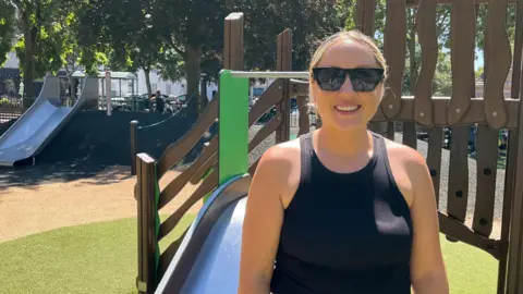 A woman wearing sunglasses stands in front of a slide in a play area and smiles at the camera.