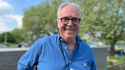Brian Mathew smiles at the camera. He is wearing a blue shirt and has grey hair and black thick-rimmed glasses