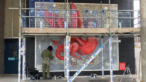 A street artist in khaki overalls spattered with paint standing under scaffolding and painting the side of a building with a large mural of a dragon with a woman's head