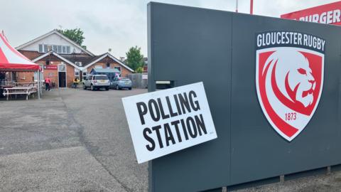 The Lion's Den with a red sign near 'polling station' sign with building and car park behind.