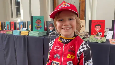 A young child in a red racing driver costume and cap grins at the camera, standing in front of a table displaying Little People, BIG DREAMS books including titles on Lewis Hamilton, Stephen Hawking, Nelson Mandela and Cristiano Ronaldo.