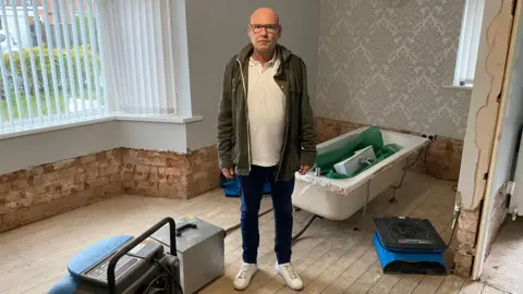 Barry Froggatt in a picture. He is standing in a living room which has been stripped and has dehumidifiers. He is making a serious face. He is a bald man wearing glasses, a white polo top and a green jacket. 