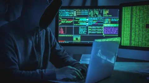 A hooded person typing into a laptop; in the background are computer screens displaying lines of code.