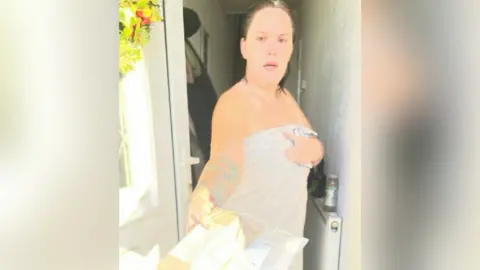 Ellie Roxanne Johnson Ellie is pictured, looking awkward, standing at her front door while holding two parcels dressed only in a grey towel.