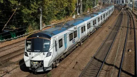A great Northern Train coming along the tracks, it is blue and silver, it is a bright sunny day and there is a tall hedge verge along the left hand side