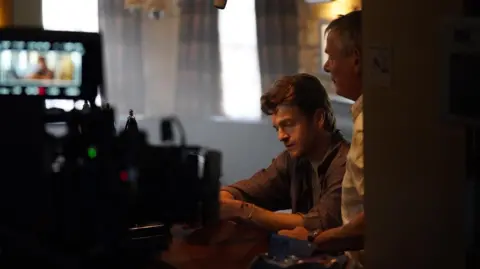 Jed Bessell Jonno Davies sits at a dimly lit bar with Martin Clunes next to him while film equipment, including a camera and overhead microphone, is positioned around them
