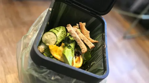 Food waste in an open bin, including bread, courgette slices and salad leaves. There is a wooden floor and the food is contained within a bag inside the bin.