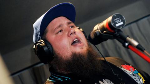 Rag'n'Bone Man wearing a baseball cap and headphones while singing into a microphone in a studio