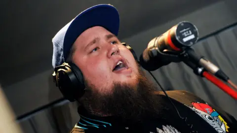 BBC Rag'n'Bone Man wearing a baseball cap and headphones while singing into a microphone in a studio