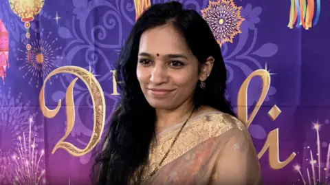 A woman standing in front of a purple banner which as lanterns as text saying Happy Diwali on it. She is wearing traditional Indian clothing.