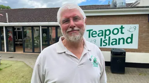 BBC/Phil Harrison A man in his sixties with white hair and a white beard and glasses looks at the camera in front of his business premises, teapot island