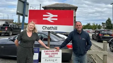 Northumberland County Council Councillors Guy Renner-Thompson and Wendy Pattison stand either side of a sign for Chathill station.