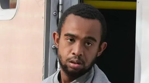 A man with short black hair and facial hair wearing a grey jumper, being led from the side of a prison van