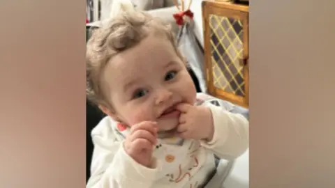 Baby with curly hair sitting in high chair - he has his finger in his mouth. He is wearing a baby grow with an elephant on it.