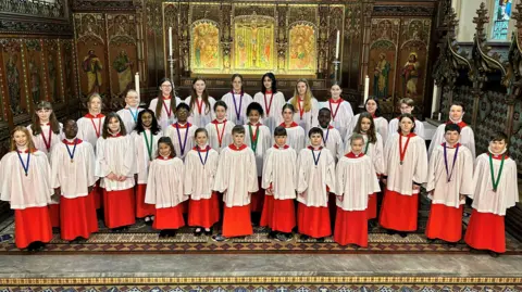 A group of young choristers wearing red and white robes. They smile at the camera as they stand within a church.