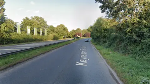 A dual carriage away which leads to a roundabout. There is a black car in the distance. The road is surrounded by trees and hedges.  There are green flags on the left side of the carriageway. 