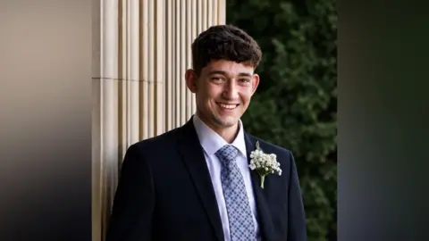A man with short brown hair smiles at the camera. He is wearing a suit with a blue tie and appears to be standing outside. 
