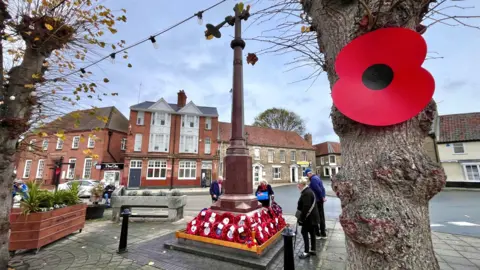 A large red poppy is pinned to a tree in the foreground. In the background a small group of people gather around a tall war memorial in a town centre. It has lots of poppy wreaths at its base.