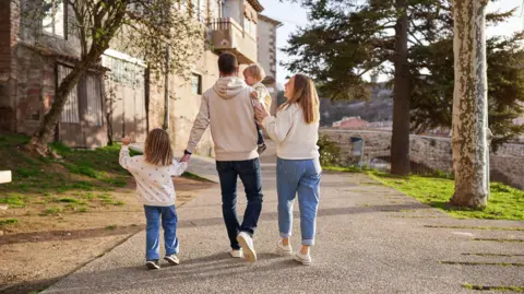 A family of four - two adults and two young children - walking down a path. There is an old building on the left and a bridge in the distance on the right. The man is wearing a cream hoodie and blue jeans with white shoes. The man is holding a child. The child to his left is wearing a white jumper with pink flowers on, blue jeans and shoes. She is holding his hand. The woman is standing to the right of a man and is wearing a white crochet jumper, light blue jeans and shoes.
