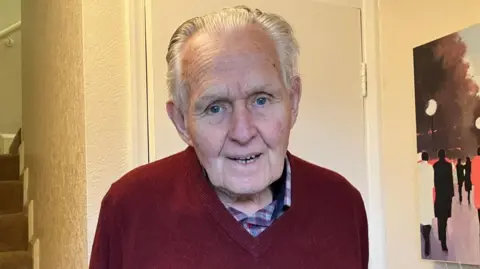 Derek, in his late 80s, stood in the doorway of his property. He has white hair combed back. He has blue eyes and is smiling. He has a chequered blue and burgundy shirt with a burgundy sweater on top.