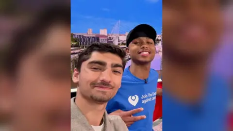 Selfie of Kush (left) has with short dark hair and Jo (right) who is wearing a cap and a blue t-shirt.