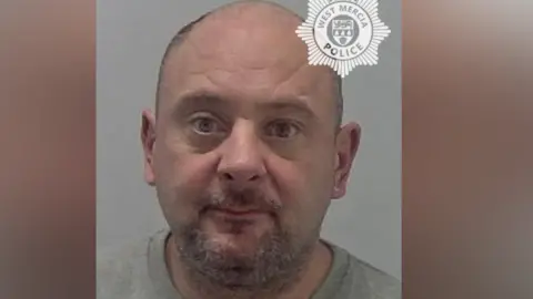 A mugshot of a man with very short hair and a stubbly beard. He is wearing a grey prison jumper and is against a neutral grey background. 