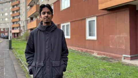 Iftikhar Ali standing in front of Westfield Court. He has dark hair and a dark goatee. He is wearing a dark jacket and is looking straight at the camera.
