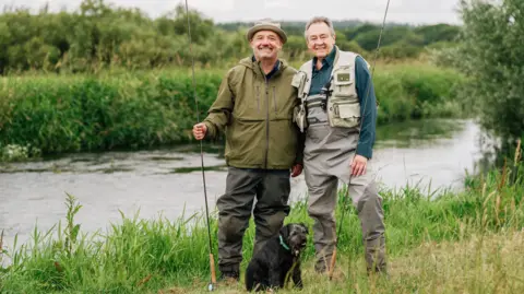 BBC/Owl Power/Sam Gibson Bob Mortimer and Paul Whitehouse pose for a photo, holding fishing lines with standing by a river, with small black dog Ted in front of them.