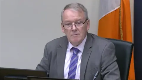 Oireachtas Brian Stanley is sitting in a committee. He's wearing black glasses and a grey suit with a white shirt and a purple tie. The Irish tricolour flag is behind him.
