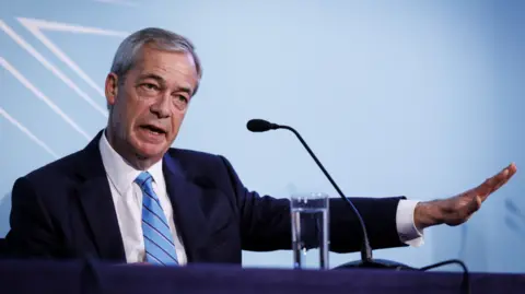 Nigel Farage wearing a dark suit and a blue tie sitting in front of a microphone and a glass of water. 