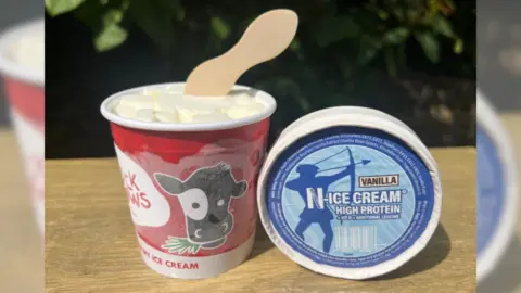 A tub of "N-ICE Cream" sat on a table, with a wooden spoon sticking out of the top of the tub.