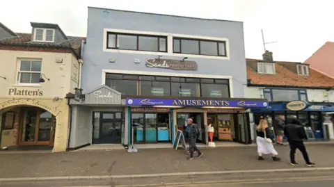 Google Street view of a seaside town high street showing Platten’s fish and chip restaurant beside Sands Restaurant and an amusement arcade. To the right is French's fish and chip shop. Several pedestrians walk along the pavement in front of a row of mixed traditional and modern shopfronts.