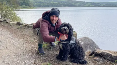 Neil Kelders is crouching down on a dirt footpath with a black, medium-sized dog. To the right of them are large rocks that line the side of a lake. Neil is wearing a black beanie hat and a dark pink/purple puffer coat.