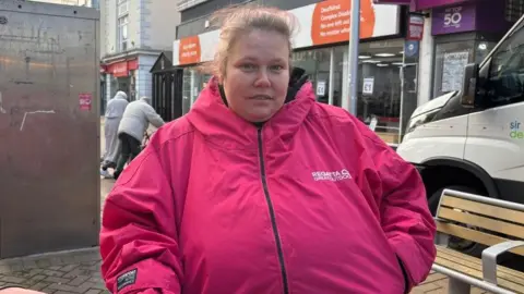A woman with light brown hair tied in a ponytail, wearing a pink coat.She is standing on the street, with shops pictured behind her.