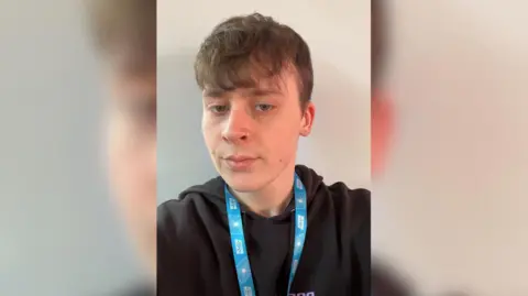 Bristol Beacon Close up of Benji Dorney wearing a black hoodie and a blue lanyard. He has brown hair and blue eyes and is stood in front of a cream background.