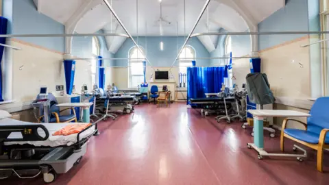 Stroud Hospitals League of Friends A hospital ward is pictured with beds either side and blue curtains. 