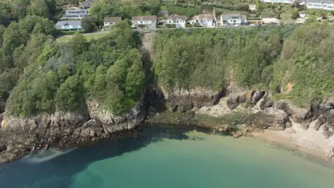 Soldier's Bay and the cliff face above it. At the top is Fort George a collection of large houses with brown roofs and white facades. Beneath the houses is a layer of greenery, with trees covering the middle half of the cliffs, above brown strips of granite getting darker as it goes closer to an azure blue sea. 