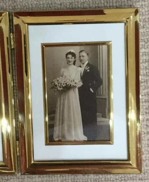 A black and white wedding photograph in a gold frame, showing a young bride in a long white gown holding a bouquet of lilies, standing beside a groom in morning dress.