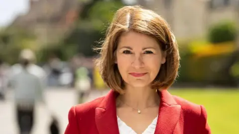 Gloucestershire County Council A headshot of Lisa Spivey, she is stood against a blurred outdoor background where people can be seen walking behind her. She has a brown bob and is wearing a red blazer, a white top, and a necklace with a pearl on it.