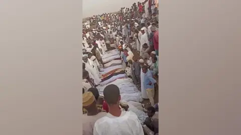 X Bodies wrapped in cloth surrounded by a large group of mourners.