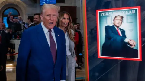 Getty Images Trump, an elderly man with blonde hair in a blue suit and purple tie, stands next to a Time magazine cover that has an image of him and the words "PERSON OF THE YEAR" on it. Melania Trump, wearing a grey pant suit, stands just behind him