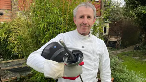 Martin Briggs, chairman of Royal Tunbridge Wells Fencing Club, dressed in protective clothing, holding a fencing mask and foil