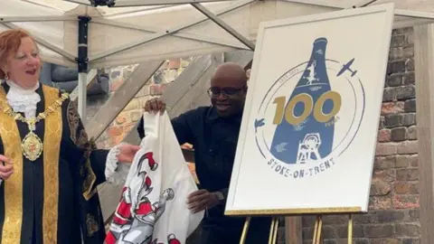 BBC A woman wearing mayoral robes and chain is seen holding a cloth and looking at a logo on an easel. There is a man wearing a black top atood to the side of the easel. On a white board is a logo with a blue bottle kiln and the number 100 in gold type.