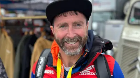 Dinny Collins wears a black cap and colourful jacket and backpack. He smiles.